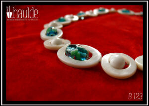 Vue en gros plan des perles du collier constitué d'ellipses de nacre blanc alternées avec des cercles de nacre blanc, à l'intérieur de chaque ellipse il y a une perle en verre de Bohème oblongue vert pâle à incrustation fleurs turquoise et rouge. A l'intérieur de chaque cercle une perle en verre blanc opaque. Le fermoir est un mousqueton flanqué de deux grosses perles en verre blanc opaque et de cercle de nacre avec une petite perle turquoise au centre