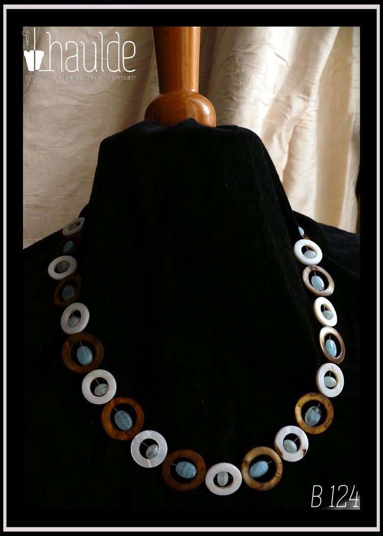 Collier composé de cercles de nacre mordoré avec en leur centre une perle ovale plate en verre bleu pâle transparent, alternés avec des cercles de nacre blanc un peu plus petits avec en leur centre un petit galet de verre bleu pâle transparent Vu autour du cou d'un mannequin de couture vêtu de velours noir
