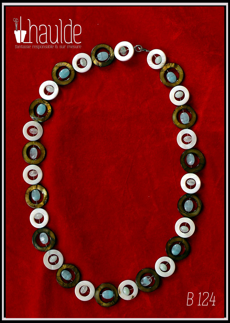 Collier composé de cercles de nacre mordoré avec en leur centre une perle ovale plate en verre bleu pâle transparent, alternés avec des cercles de nacre blanc un peu plus petits avec en leur centre un petit galet de verre bleu pâle transparent Vu de dessus sur du velours rouge vif