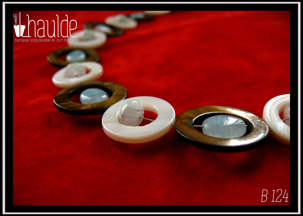 Vue en gros plan des perles d'un collier composé de cercles de nacre mordoré avec en leur centre une perle ovale plate en verre bleu pâle transparent, alternés avec des cercles de nacre blanc un peu plus petits avec en leur centre un petit galet de verre bleu pâle transparent