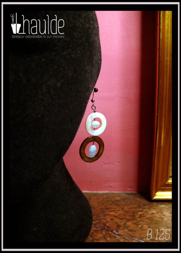 Boucles d'oreilles montées sur crochet composées d'un cercle de nacre mordoré avec en son centre une perle ovale plate en verre bleu pâle transparent surmonté d'un cercle de nacre blanc plus petit avec en son centre un galet de verre bleu pâle Vues sur une tête de mannequin en velours noir