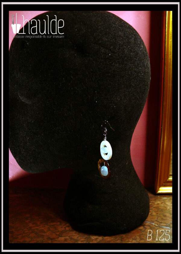 Boucles d'oreilles montées sur crochet composées d'un cercle de nacre mordoré avec en son centre une perle ovale plate en verre bleu pâle transparent surmonté d'un cercle de nacre blanc plus petit avec en son centre un galet de verre bleu pâle Vu de profil sur une tête de mannequin en velours noir