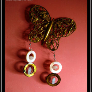 Boucles d'oreilles montées sur crochets, composées d'un cercle de nacre mordoré avec en son centre une perle ovale plate en verre bleu pâle, surmontée d'un cercle de nacre blanc plus petit avec en son centre un galet en verre bleu pâle La paire vue suspendue à un papillon de dentelle de métal doré
