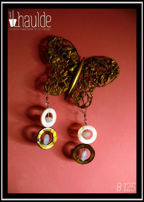 Boucles d'oreilles montées sur crochets, composées d'un cercle de nacre mordoré avec en son centre une perle ovale plate en verre bleu pâle, surmontée d'un cercle de nacre blanc plus petit avec en son centre un galet en verre bleu pâle La paire vue suspendue à un papillon de dentelle de métal doré