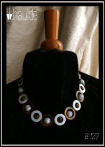 Collier fermé par un mousqueton, composé de cercles de nacre blanc avec en leur centre une perle de jaspe unakite (chamarré vert kaki et marron roux) alternés avec des cercles de nacre mordorés un peu plus grand avec en leur centre une perle de verre blanc opaque rayé de fines lignes marrons Vu autour du cou d'un mannequin de couture vêtu de velours noir