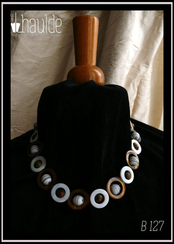 Collier fermé par un mousqueton, composé de cercles de nacre blanc avec en leur centre une perle de jaspe unakite (chamarré vert kaki et marron roux) alternés avec des cercles de nacre mordorés un peu plus grand avec en leur centre une perle de verre blanc opaque rayé de fines lignes marrons Vu autour du cou d'un mannequin de couture vêtu de velours noir