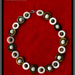 Collier fermé par un mousqueton, composé de cercles de nacre blanc avec en leur centre une perle de jaspe unakite (chamarré vert kaki et marron roux) alternés avec des cercles de nacre mordorés un peu plus grand avec en leur centre une perle de verre blanc opaque rayé de fines lignes marrons Vue de dessus sur du velours rouge vif