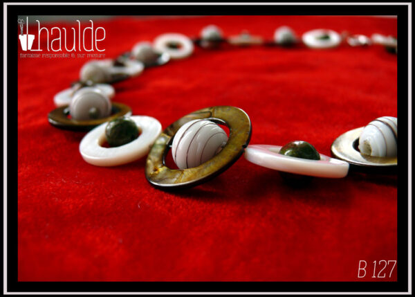 Vue en gros plan des perles composant un collier fermé par un mousqueton, composé de cercles de nacre blanc avec en leur centre une perle de jaspe unakite (chamarré vert kaki et marron roux) alternés avec des cercles de nacre mordorés un peu plus grand avec en leur centre une perle de verre blanc opaque rayé de fines lignes marrons
