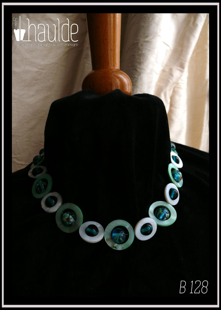 Collier, fermoir à mousqueton, composé de cercles de nacre vert pâle avec en leur centre une perle ronde en verre de Bohème turquoise pâle à inclusion fleurs vert blanc et orange, alternés avec des cercles de nacre blanc plus petits avec en leur centre une perle de verre transparent bleu paon Vu autour du cou d'un mannequin de couture vêtu de velours noir