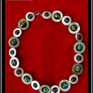 Collier, fermoir à mousqueton, composé de cercles de nacre vert pâle avec en leur centre une perle ronde en verre de Bohème turquoise pâle à inclusion fleurs vert blanc et orange, alternés avec des cercles de nacre blanc plus petits avec en leur centre une perle de verre transparent bleu paon Vu de dessus sur du velours rouge vif