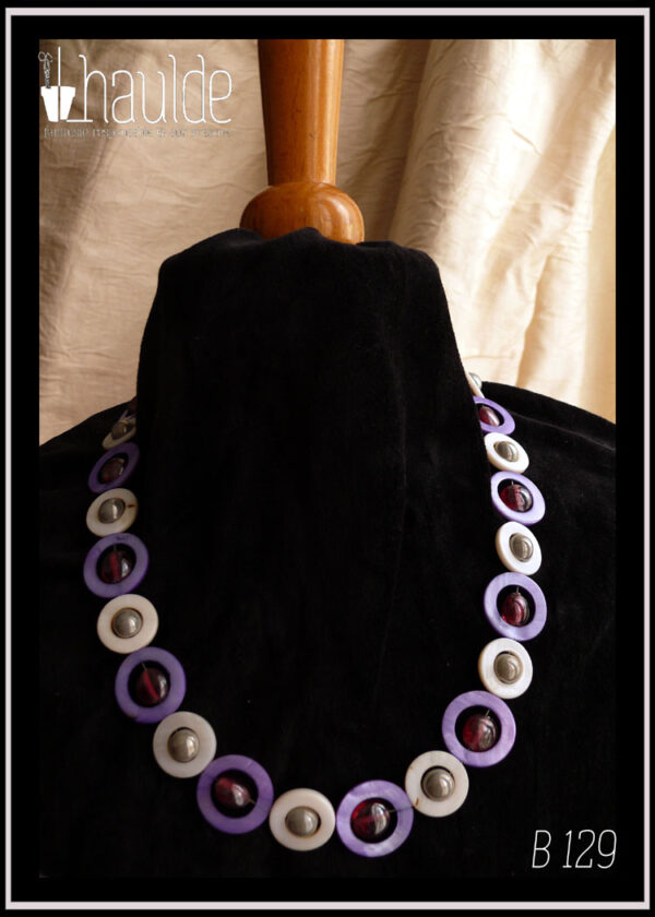 Collier, fermoir à mousqueton, composé de cercles de nacre violets avec en leur centre une perle plate en verre rose transparent et métallisé, alternés avec des cercles de nacre blanc plus petits avec en leur centre une perle en verre gris opaque et nacré Vu autour du cou d'un mannequin de couture vêtu de velours noir