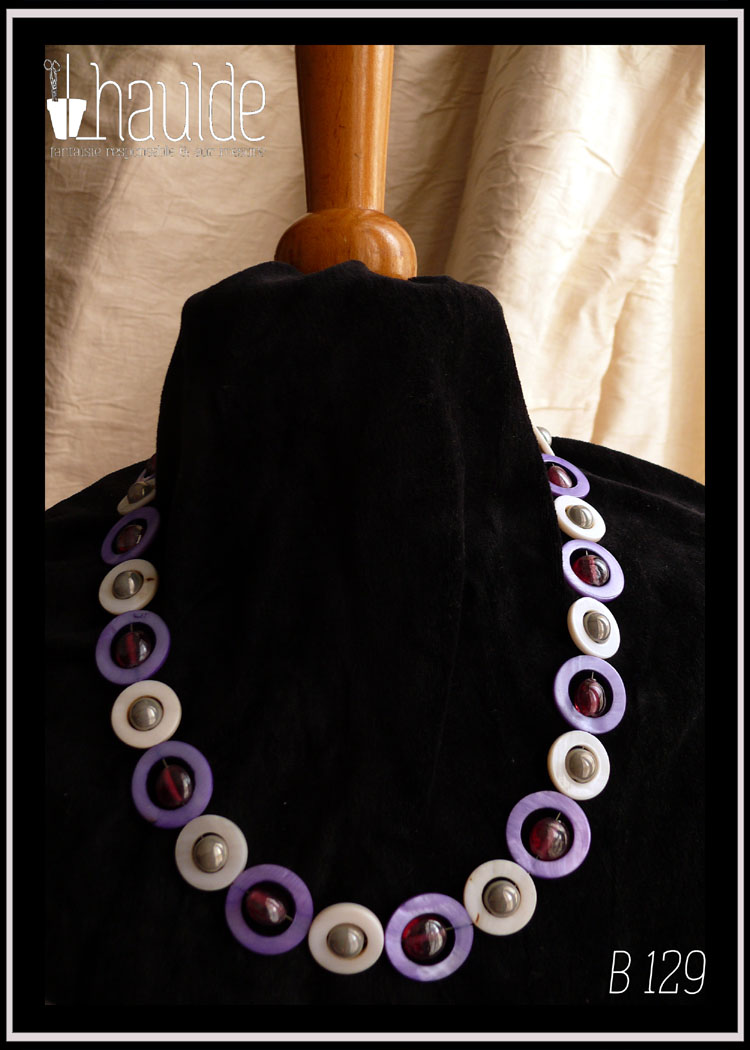 Collier, fermoir à mousqueton, composé de cercles de nacre violets avec en leur centre une perle plate en verre rose transparent et métallisé, alternés avec des cercles de nacre blanc plus petits avec en leur centre une perle en verre gris opaque et nacré Vu autour du cou d'un mannequin de couture vêtu de velours noir