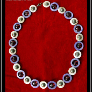 Collier, fermoir à mousqueton, composé de cercles de nacre violets avec en leur centre une perle plate en verre rose transparent et métallisé, alternés avec des cercles de nacre blanc plus petits avec en leur centre une perle en verre gris opaque et nacré Vu de dessus sur du velours rouge vif