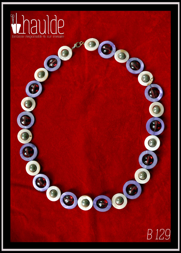 Collier, fermoir à mousqueton, composé de cercles de nacre violets avec en leur centre une perle plate en verre rose transparent et métallisé, alternés avec des cercles de nacre blanc plus petits avec en leur centre une perle en verre gris opaque et nacré Vu de dessus sur du velours rouge vif