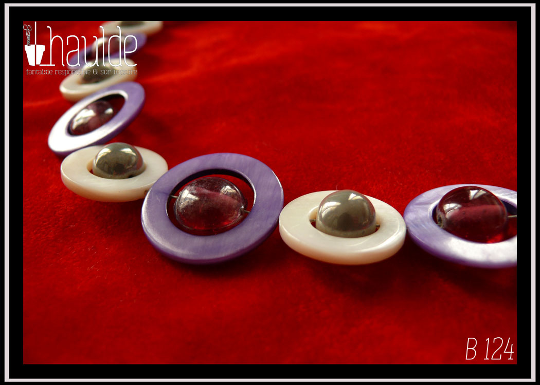 Vue en gros plan des perles du collier, fermoir à mousqueton, composé de cercles de nacre violets avec en leur centre une perle plate en verre rose transparent et métallisé, alternés avec des cercles de nacre blanc plus petits avec en leur centre une perle en verre gris opaque et nacré