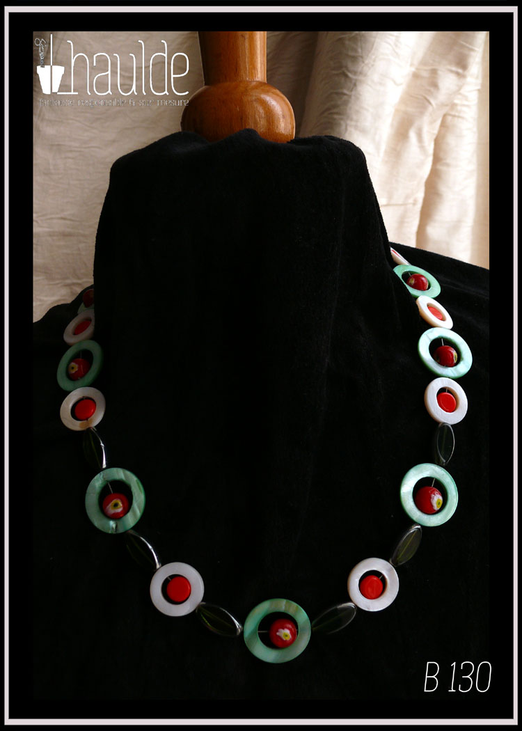 Collier, fermoir à mousqueton, composé de cercles de nacre vert avec en leur centre une perle de verre de Bohème rouge à inclusion fleur jaune et blanc, alternés avec des cercles de nacre blancs plus petits avec en leur centre un palet de verre rouge vif. Au centre du collier, devant, les cercles de nacre sont séparés par des perles de verre vert transparent, tranche acier en forme de feuille Vu autour du cou d'un mannequin de couture vêtu de velours noir