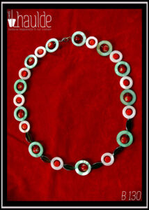 Collier, fermoir à mousqueton, composé de cercles de nacre vert avec en leur centre une perle de verre de Bohème rouge à inclusion fleur jaune et blanc, alternés avec des cercles de nacre blancs plus petits avec en leur centre un palet de verre rouge vif. Au centre du collier, devant, les cercles de nacre sont séparés par des perles de verre vert transparent, tranche acier en forme de feuille Vu de dessus sur du velours rouge vif