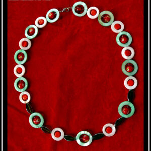 Collier, fermoir à mousqueton, composé de cercles de nacre vert avec en leur centre une perle de verre de Bohème rouge à inclusion fleur jaune et blanc, alternés avec des cercles de nacre blancs plus petits avec en leur centre un palet de verre rouge vif. Au centre du collier, devant, les cercles de nacre sont séparés par des perles de verre vert transparent, tranche acier en forme de feuille Vu de dessus sur du velours rouge vif