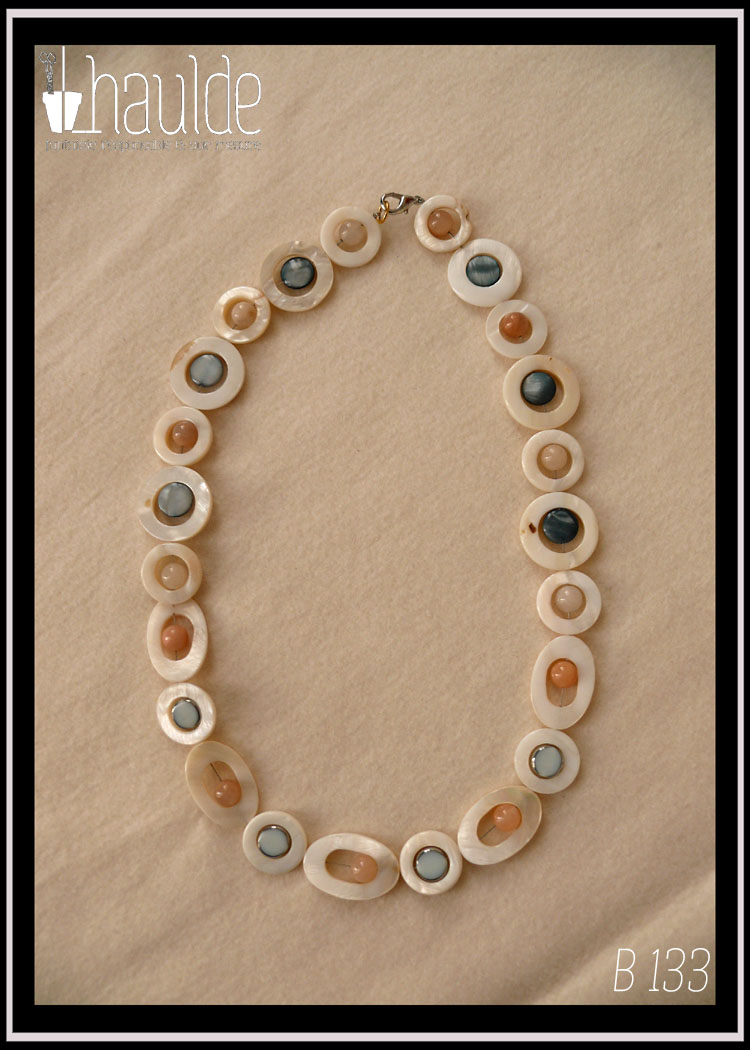 Collier composé de cercles de nacre blanc avec un palet de nacre bleu en leur centre, alterné avec des cercles de nacre blanc plus petits avec une perle d'aventurine rose pâle en leur centre. Au milieu du collier les petits cercles de nacre ont une perle en verre bleu pâle plate à tranche d'acier en leur centre et sont alternés avec des ellipses de nacre blanc avec une perle d'aventurine rose en leur centre Vu de dessus posé sur de la polaire blanche