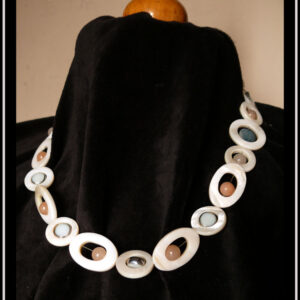 Collier composé de cercles de nacre blanc avec un palet de nacre bleu en leur centre, alterné avec des cercles de nacre blanc plus petits avec une perle d'aventurine rose pâle en leur centre. Au milieu du collier les petits cercles de nacre ont une perle en verre bleu pâle plate à tranche d'acier en leur centre et sont alternés avec des ellipses de nacre blanc avec une perle d'aventurine rose en leur centre Vu porté par un mannequin de couture vêtu de velours noir