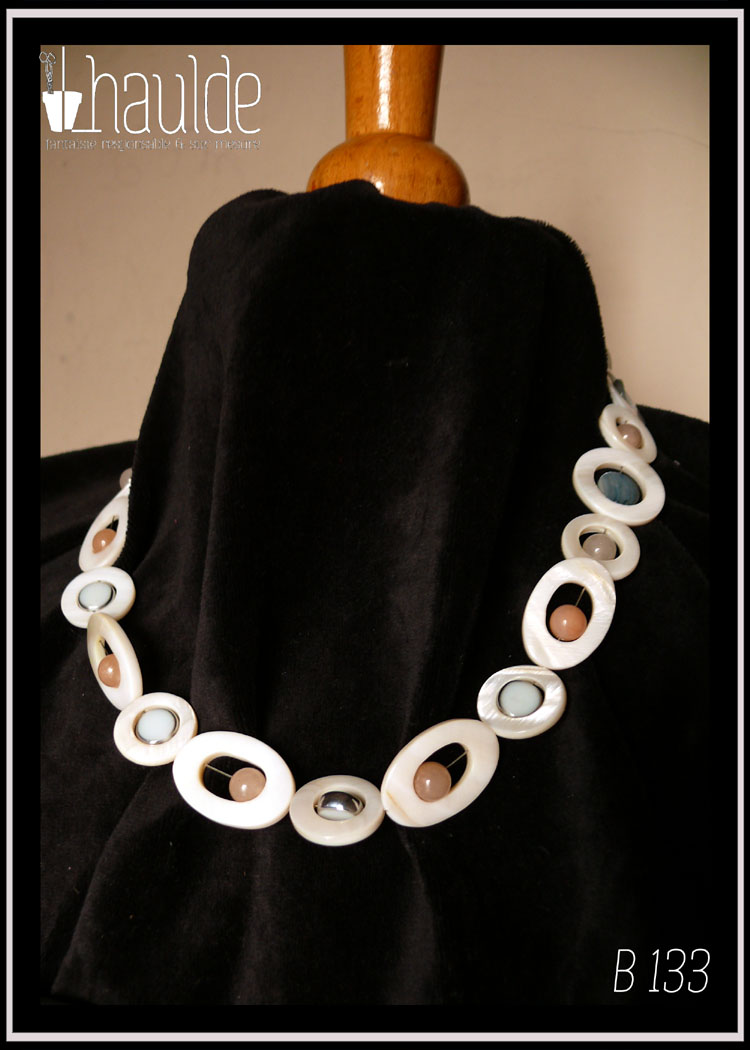 Collier composé de cercles de nacre blanc avec un palet de nacre bleu en leur centre, alterné avec des cercles de nacre blanc plus petits avec une perle d'aventurine rose pâle en leur centre. Au milieu du collier les petits cercles de nacre ont une perle en verre bleu pâle plate à tranche d'acier en leur centre et sont alternés avec des ellipses de nacre blanc avec une perle d'aventurine rose en leur centre Vu porté par un mannequin de couture vêtu de velours noir