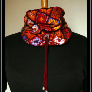 cache-col montant en jersey imprimé "granny squares", des carrés de crochets multicolores, dominante rouge. Doublure en velours rouge bordeaux, cordon rouge bordeaux avec des clochettes pour protéger les bouts et un serre-cordon transparent. Vu côté jersey imprimé sur un mannequin de couture