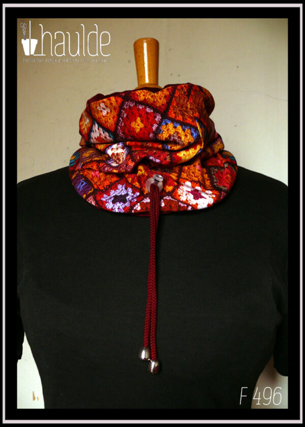 cache-col montant en jersey imprimé "granny squares", des carrés de crochets multicolores, dominante rouge. Doublure en velours rouge bordeaux, cordon rouge bordeaux avec des clochettes pour protéger les bouts et un serre-cordon transparent. Vu côté jersey imprimé sur un mannequin de couture
