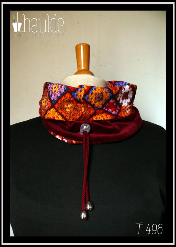 cache-col montant en jersey imprimé "granny squares", des carrés de crochets multicolores, dominante rouge. Doublure en velours rouge bordeaux, cordon rouge bordeaux avec des clochettes pour protéger les bouts et un serre-cordon transparent. Vu avec le jersey replié sur le velours sur un mannequin de couture