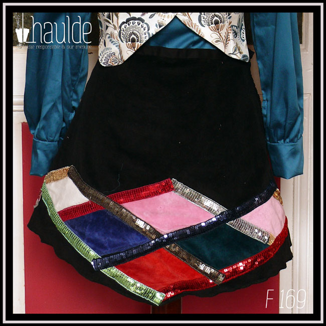 mini jupe trapèze en velours milleraies noir, losanges de velours rouge, rose pâle, blanc, bleu roi et bleu pétrole encadré de galons de sequins doré, vert, argent, rouge et rouge-rose Vue de face sur un mannequin de couture qui porte également un chemisier en satin bleu paon dont on voit le bout des manches et un boléro en toile blanche imprimée