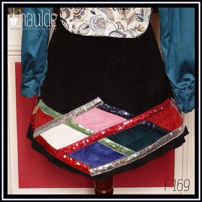 mini jupe trapèze en velours milleraies noir, losanges de velours rouge, rose pâle, blanc, bleu roi et bleu pétrole encadré de galons de sequins doré, vert, argent, rouge et rouge-rose Vue de dos sur un mannequin de couture qui porte également un chemisier en satin bleu paon dont on voit le bout des manches et un boléro en toile blanche imprimée