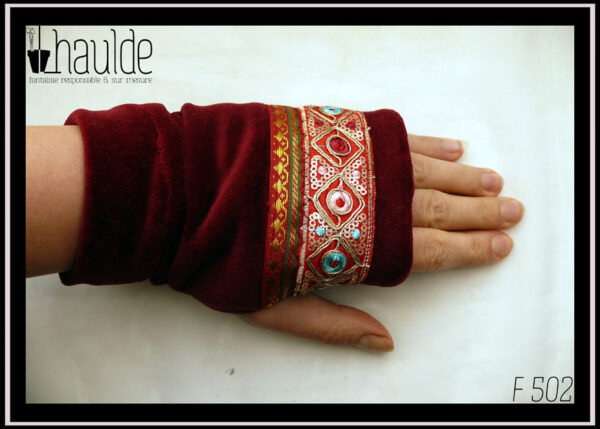 Mitaine en velours rouge bordeaux, garnie de deux galons brodés, un large brodé de losanges dorés avec au centre un cercle soit rouge bordeaux, soit bleu pâle, soit rose pâle et garni de petits sequins circulaires dorés, le deuxième plus étroit en satin rouge bordeaux brodé de motifs géométriques (pointes de flèche) dorés et verts foncés Les galons sont placés entre les phalanges et le milieu de l'ouverture pour le pouce Vue portée côté dos de la main