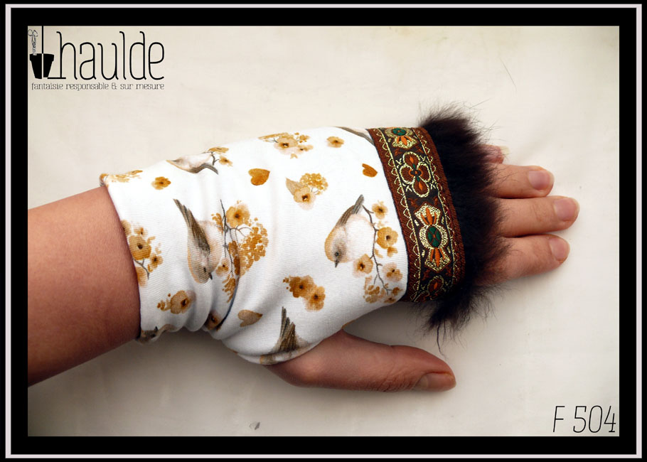 Paire de mitaines en jersey blanc imprimé petits oiseaux et petites fleurs dans les tons gris, rosés et ocres Elles sont garnies à une extrémité d'un galon marron brodé de motifs abstraits vaguement floraux dorés oranges et verts avec des la fourrure marron Vue de dessus d'une mitaine portée, la fourrure en haut côté droit, les oiseaux à l'envers (le galon est censé être côté manchette mais les mitaines sont trop étroites pour mes mains dans ce sens là)