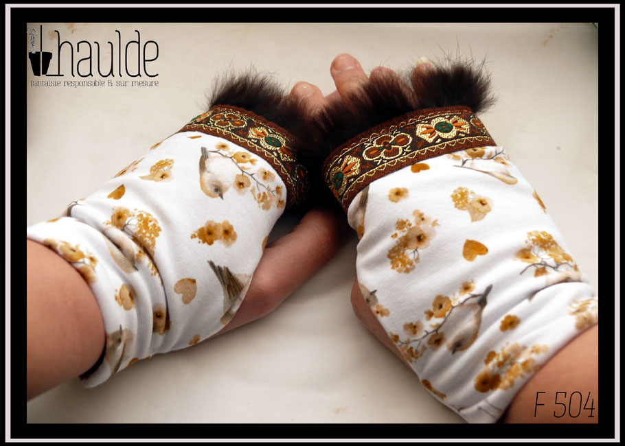 Paire de mitaines en jersey blanc imprimé petits oiseaux et petites fleurs dans les tons gris, rosés et ocres Elles sont garnies à une extrémité d'un galon marron brodé de motifs abstraits vaguement floraux dorés oranges et verts avec des la fourrure marron Vue de dessus de la paire portée, la fourrure en haut côté droit, les oiseaux à l'envers (le galon est censé être côté manchette mais les mitaines sont trop étroites pour mes mains dans ce sens là)