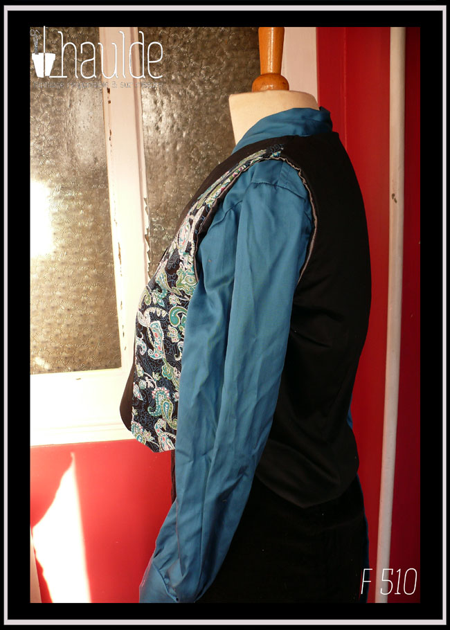 Boléro en satin noir et tissu imprimé motifs paisley bleu et turquoise sur fond noir. Le centre du boléro et le dos sont en satin noir, les côtés en tissu imprimé Vu de profil sur un mannequin de couture qui porte également un chemisier de satin bleu paon et une jupe en velours noir