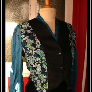 Boléro en satin noir et tissu imprimé motifs paisley bleu et turquoise sur fond noir. Le centre du boléro et le dos sont en satin noir, les côtés en tissu imprimé Vu de trois quarts face sur un mannequin de couture qui porte également un chemisier de satin bleu paon et une jupe en velours noir