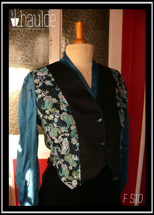 Boléro en satin noir et tissu imprimé motifs paisley bleu et turquoise sur fond noir. Le centre du boléro et le dos sont en satin noir, les côtés en tissu imprimé Vu de trois quarts face sur un mannequin de couture qui porte également un chemisier de satin bleu paon et une jupe en velours noir