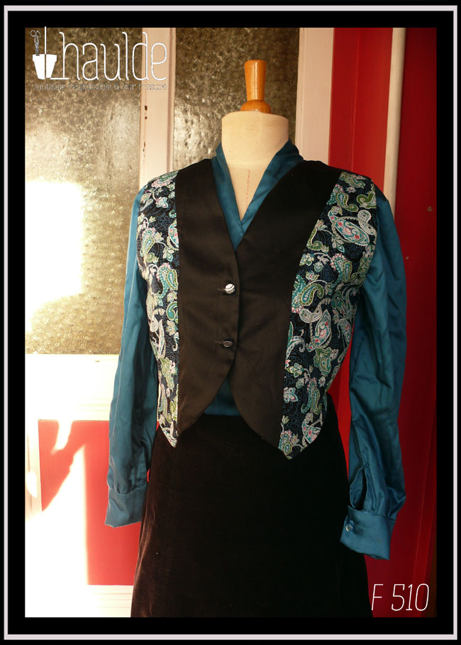 Boléro en satin noir et tissu imprimé motifs paisley bleu et turquoise sur fond noir. Le centre du boléro et le dos sont en satin noir, les côtés en tissu imprimé Fermé par deux boutons à pied translucides bleu et doré Vu de face sur un mannequin de couture qui porte également un chemisier de satin bleu paon et une jupe en velours noir