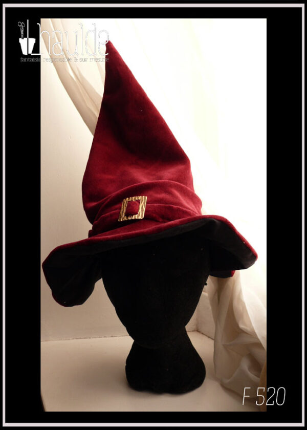 Chapeau à long bout pointu en velours rouge bordeaux avec un bandeau rehaussé d'une boucle de ceinture dorée martelée Il se tient à la verticale grâce à la doublure en sergé noir que l'on devine sous le rebord Vu porté par une tête de mannequin sur un rebord de fenêtre très blanc