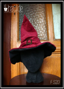 Chapeau à long bout pointu en velours rouge bordeaux avec un bandeau rehaussé d'une boucle de ceinture dorée martelée Il se tient à la verticale grâce à la doublure en sergé noir que l'on devine sous le rebord Vu porté par une tête de mannequin