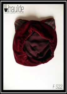 Bonnet à oreilles de chat en velours rouge bordeaux, intérieur des oreilles en écossais gris, bordeaux et rose. La doublure est en tencel à petits carreaux violets sur fond mauve que l'on voit ici puisque le bonnet est posé sur une surface blanche