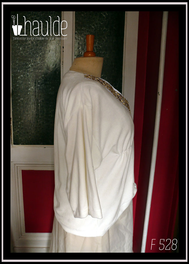 Haut manches kimono trois quarts en velours blanc écru, encolure en V profonde soulignée par un galon brodé de motifs floraux et végétaux dorés. Le devant est plus court (remonte juste en dessous du nombril) le dos retombe sur les hanches Vu de profil sur un mannequin de couture