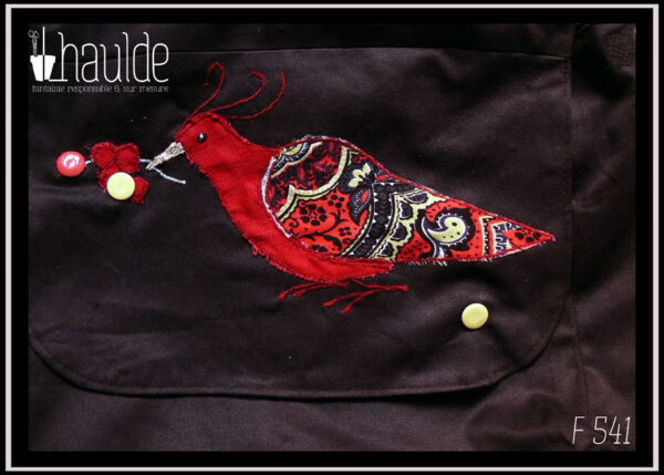 Sac à rabat et bandoulière en sergé noir, avec brodé sur le rabat un appliqué en forme d'oiseau, corps en lin rouge, aile en tissu imprimé paisley rouge L'oiseau tient dans son bec une grappe de petites baies rouges, parmi lesquelles se trouve deux boutons pressions, un rouge et un vert pâle. Un autre bouton vert pâle en bas du rabat complète le système de fermeture