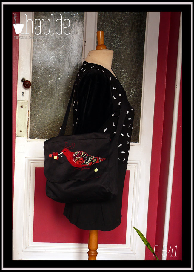 Sac à rabat et bandoulière en sergé noir, avec brodé sur le rabat un appliqué en forme d'oiseau, corps en lin rouge, aile en tissu imprimé paisley rouge L'oiseau tient dans son bec une grappe de petites baies rouges, parmi lesquelles se trouve deux boutons pressions, un rouge et un vert pâle. Un autre bouton vert pâle en bas du rabat complète le système de fermeture Vu porté par un mannequin de couture