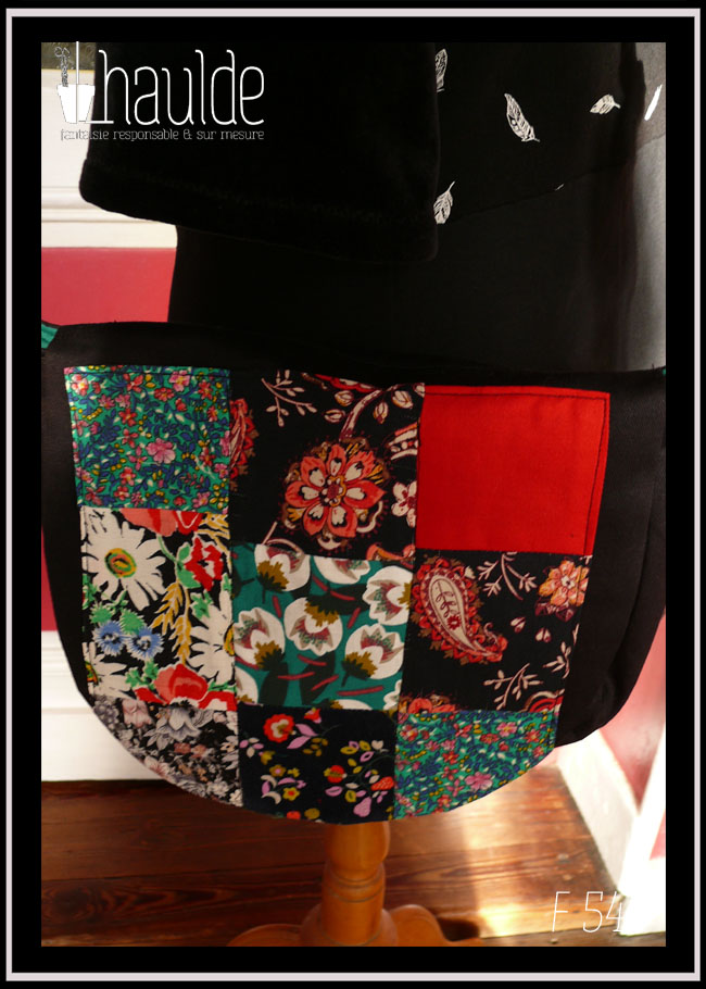 Sac à rabat en sergé noir, bords arrondis, bandoulière réglable sangle noire fleurie, avec sur le devant et sur le rabat un patchwork de carrés aux motifs fleuris, tons rouges et verts dominants. Vu avec le rabat rabattu