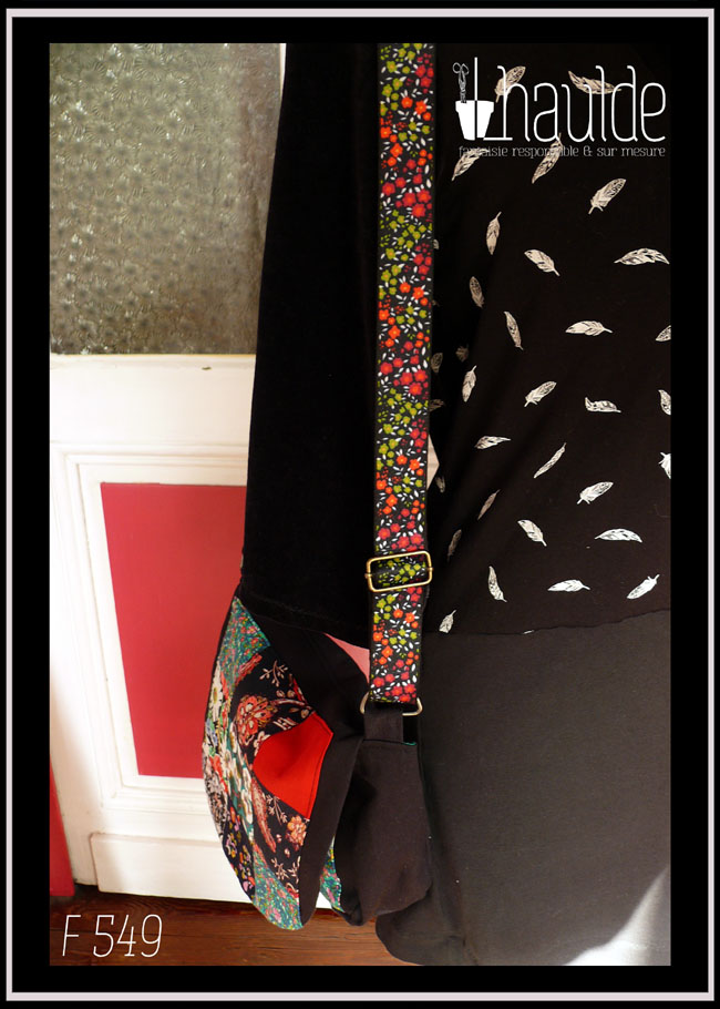 Sac à rabat en sergé noir, bords arrondis, bandoulière réglable sangle noire fleurie, avec sur le devant et sur le rabat un patchwork de carrés aux motifs fleuris, tons rouges et verts dominants. Vu de profil pour voir la sangle fleurie