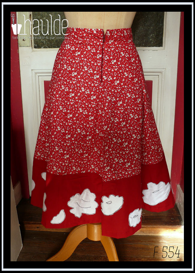 Jupe mi longue à pans amples ajustés à la taille en tissu rouge foncé imprimé petites fleurs blanches. Le bas de la jupe est une large bande de tissu rouge foncé avec des appliqués en satin blanc qui reprennent les motifs floraux du tissus imprimé Vue de dos sur un mannequin de couture