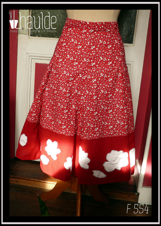 Jupe mi longue à pans amples ajustés à la taille en tissu rouge foncé imprimé petites fleurs blanches. Le bas de la jupe est une large bande de tissu rouge foncé avec des appliqués en satin blanc qui reprennent les motifs floraux du tissus imprimé Vue de face sur un mannequin de couture