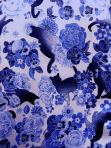 tissu fond bleu pâle imprimé chats noirs qui se baladent entre des bouquets de roses et de pensées bleues cernées de blanc, ce qui crée un effet comme si elles étaient fantomatiques
