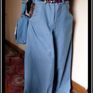 Jean ample bleu clair dont l'intérieur des poches est garni de tissu imprimé prune à motifs dorés. Le bord des poches est souligné par des oeillets dorés Vu sur un mannequin de couture avec une ceinture fine en simili cuir violet, un haut et un sac en jean