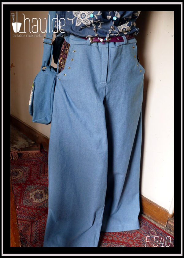 Jean ample bleu clair dont l'intérieur des poches est garni de tissu imprimé prune à motifs dorés. Le bord des poches est souligné par des oeillets dorés Vu sur un mannequin de couture avec une ceinture fine en simili cuir violet, un haut et un sac en jean